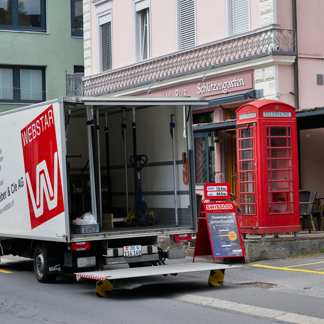 A moving truck with an open rear cargo area parked on a street in front of a building featuring pastel pink and green facades, with a decorative balcony and large windows. The truck displays the Webst