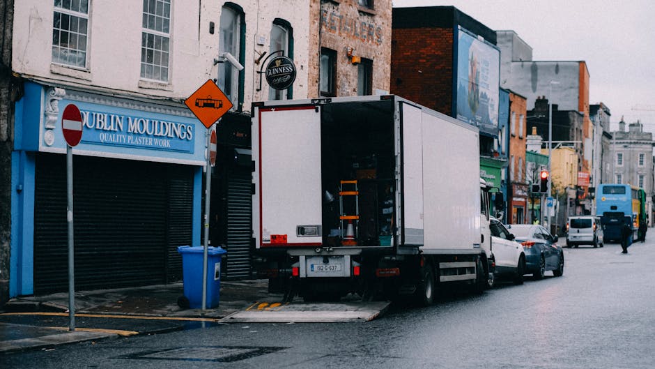 A moving truck with an open rear cargo area parked on a street in front of a building featuring pastel pink and green facades, with a decorative balcony and large windows. The truck displays the Webst